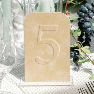 May include: A beige table number five, displayed on a clear acrylic stand. The number is embossed on the rectangular sign. The background includes a lit candle, grapes, and a fork, suggesting a wedding or event setting.