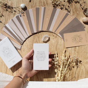Könnte beinhalten: Ein Kartenspiel "PROSPERITY" mit einer weißen Box und einer gehaltenen Karte. Die Karten sind auf einer Holzoberfläche mit getrockneten Blumen und Kristallen ausgebreitet. Die Karte in der Hand trägt den Text: "Wo biete ich eine andere Perspektive an?"