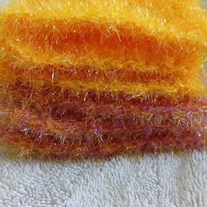 May include: A stack of handmade, textured scrubbies in shades of orange and pink. The scrubbies are made of a fluffy, yarn-like material and are stacked on top of each other. The scrubbies are sitting on a white, textured surface.