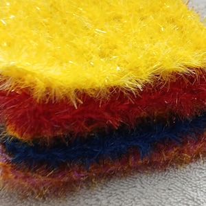 Puede incluir: Una pila de estropajos coloridos y texturizados en amarillo, rojo y azul. Los estropajos tienen un aspecto esponjoso y desgreñado, y probablemente se utilizan para limpiar o exfoliar. Los colores son vibrantes y la textura es llamativa.