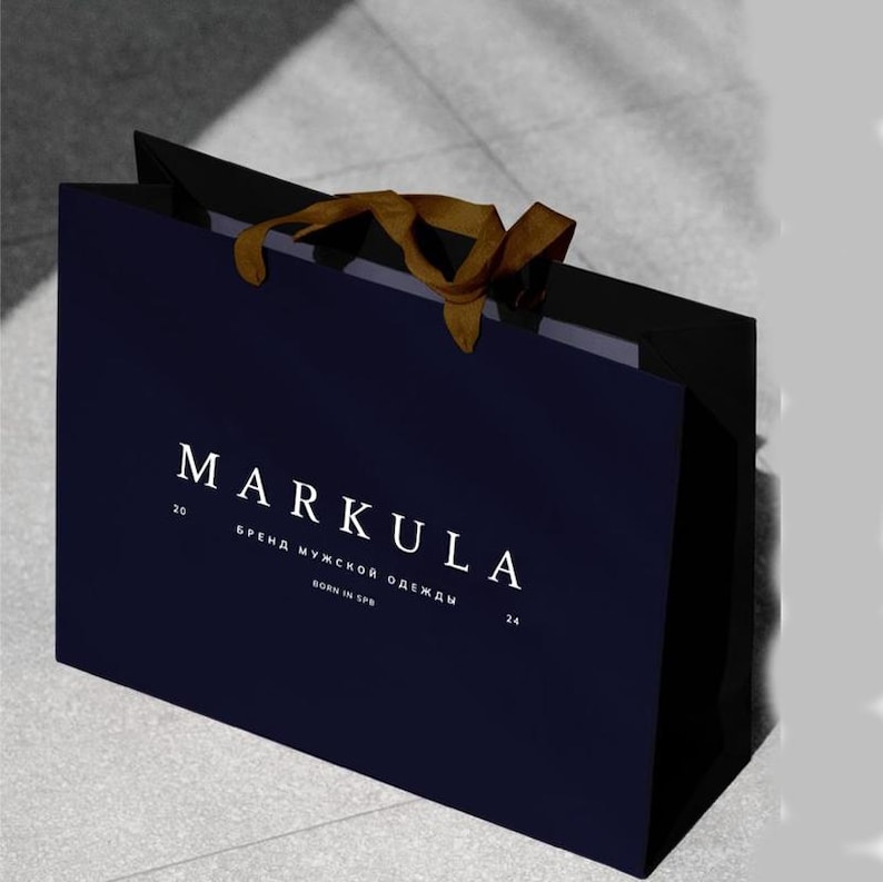 May include: A navy blue shopping bag with the brand name "MARKULA" in white lettering. The bag has a gold ribbon handle and additional text in a smaller font. The bag is likely for clothing or accessories.