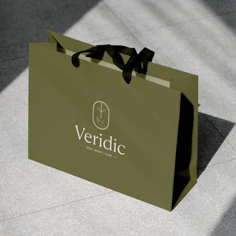 May include: Olive green shopping bag with black ribbon handles. The bag features the white text "Veridic" with the tagline "SKIN | BODY | HAIR" and a logo of a hand holding a plant. The bag is sitting on a gray surface.