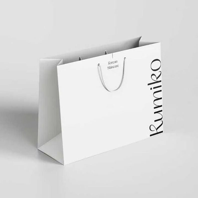 K&ouml;nnte beinhalten: Eine wei&szlig;e Papiereinkaufstasche mit dem Wort "Rumiko" in schwarzer Schrift vertikal auf der rechten Seite. Die Tasche hat einen silbernen Kordelgriff und die Worte "Korean Skincare" sind &uuml;ber dem Griff aufgedruckt.
