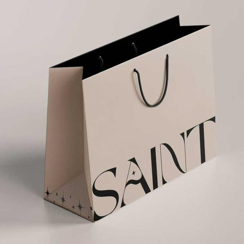K&ouml;nnte beinhalten: Eine beige Einkaufstasche mit dem Wort "SAINT" in gro&szlig;en schwarzen Buchstaben aufgedruckt. Die Tasche hat ein schwarzes Innenfutter und schwarze Kordelgriffe. Die untere Ecke zeigt ein Muster aus kleinen blauen Sternformen.