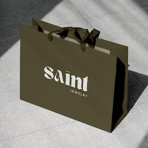 May include: An olive green shopping bag with the text "Saint Jewelry" in white. The bag has a rectangular shape with a folded top and dark brown handles. The bag is sitting on a gray tiled surface.