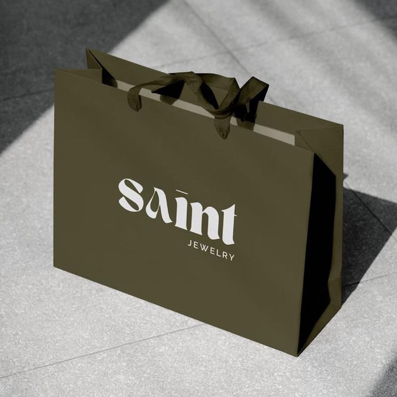 May include: An olive green shopping bag with the text "Saint Jewelry" in white. The bag has a rectangular shape with a folded top and dark brown handles. The bag is sitting on a gray tiled surface.