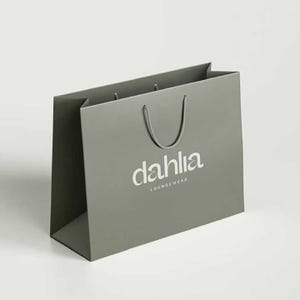K&ouml;nnte beinhalten: Eine graue Einkaufstasche mit dem Wort "dahlia" in wei&szlig;er Schrift und dem Wort "loungewear" darunter. Die Tasche hat eine rechteckige Form mit gefaltetem Oberteil und einem grauen Kordelgriff.