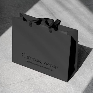 May include: A black shopping bag with a matte finish, featuring the text "Chernova decor" in a delicate font. The bag has black ribbon handles and is set against a gray tiled surface. The text "МАСТЕРСКАЯ ЮЛИИ ЧЕРНОВОЙ" is below the main text.