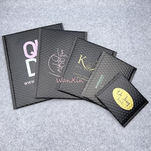 Könnte beinhalten: Eine Ansammlung schwarzer Luftpolstertaschen mit verschiedenen Logos und Texten in Rosa, Gold und Weiß. Die Logos sind auf den Taschen aufgedruckt. Die Taschen sind in verschiedenen Größen angeordnet und überlappen sich leicht.