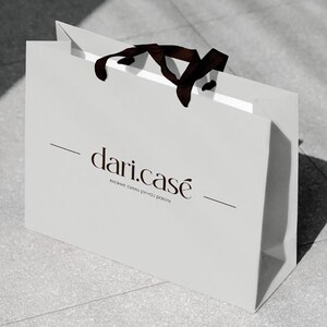 May include: A white shopping bag with a dark brown ribbon handle. The bag features the text "dari.case" in a stylish font, with additional text below. The bag is sitting on a light gray surface.