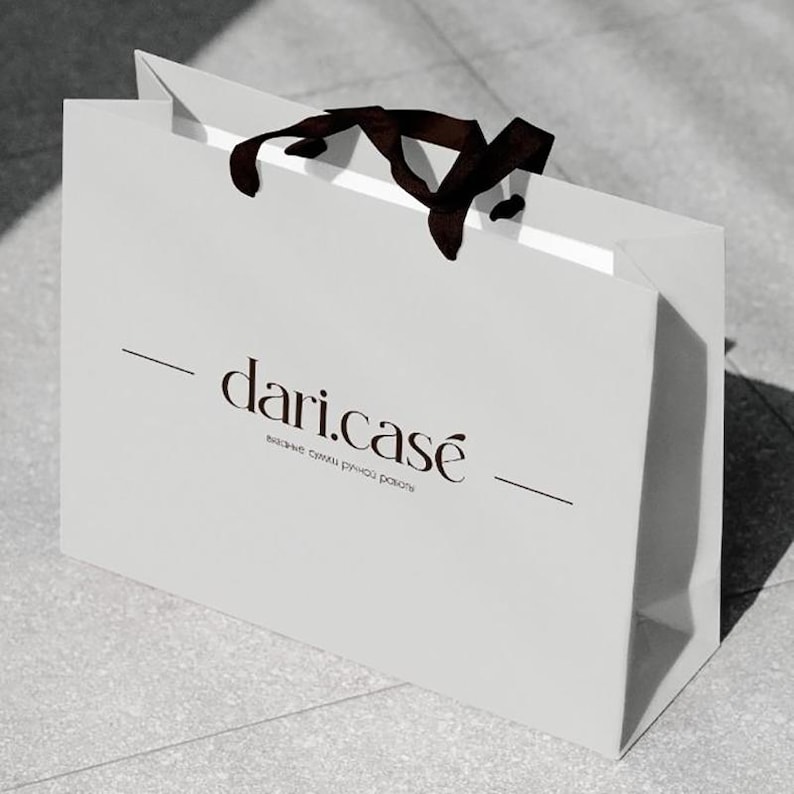 May include: A white shopping bag with a dark brown ribbon handle. The bag features the text "dari.case" in a stylish font, with additional text below. The bag is sitting on a light gray surface.