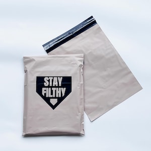 May include: Two beige poly mailer shipping bags. One bag has the text "STAY FILTHY" in white block letters inside a black baseball home plate graphic. The other bag is plain. Both bags have a black adhesive strip.