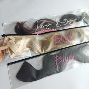 May include: Three clear plastic bags with black zippers, each containing hair extensions. The top bag has black hair with pink highlights, the middle bag has blonde hair, and the bottom bag has dark hair. The bags have the text "Lasaya Boutique" and "Blush Extensions".