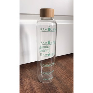 May include: A clear glass water bottle with a bamboo lid and a green and white sticker with the text "8 AM 2 PM", "9 AM 3 PM", "10 AM 4 PM", "11 AM 5 PM", "PM 6 PM", and "REFILL".