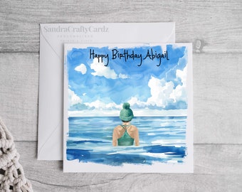 May include: A personalised birthday card featuring a watercolour illustration of a person wearing a green beanie and swimsuit standing in the sea. The card reads "Happy Birthday Abigail".