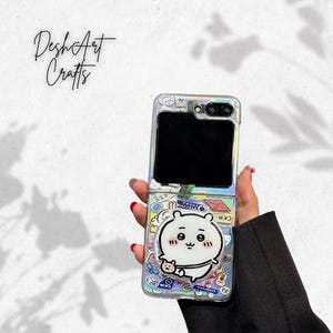 Könnte beinhalten: Eine transparente Handyhülle mit einem Cartoon-Charakter-Design. Die offene Hülle zeigt den Bildschirm des Telefons. Das Design umfasst einen weißen Cartoon-Charakter mit einem niedlichen Ausdruck, umgeben von bunten Aufklebern.