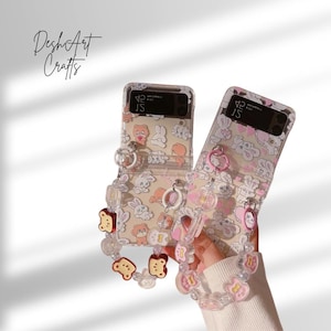 May include: Two clear phone cases featuring a repeating pattern of cartoon bears and bunnies. Each case has a matching wrist strap with bear and heart charms. The text "DeshArt Crafts" is in the upper left corner.