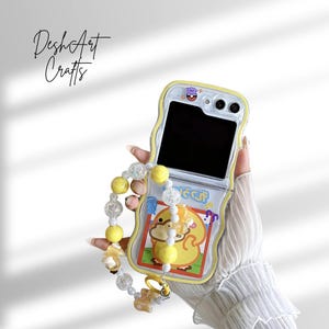 May include: A yellow and clear phone case with a cartoon duck design. The case has a beaded strap with yellow and clear beads. The text "DeshArt Crafts" is written in cursive on the top left.
