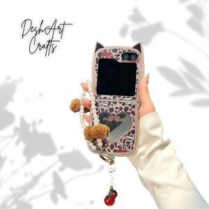 May include: A pink and brown patterned phone case with cat ears, a heart-shaped mirror, and a pearl and pom-pom wrist strap. The case has a cherry charm and the text "Desh Art Crafts".