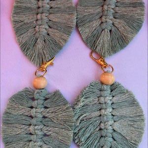 May include: Four macrame leaf-shaped earrings in shades of green. Each earring features a central woven design, a wooden bead, and a gold-colored clasp. The earrings are displayed on a light purple surface.