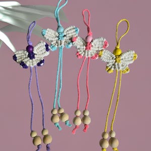 May include: Four macrame butterfly ornaments featuring off-white knotted cord wings with colored accents in purple, light blue, pink, and yellow. Each butterfly includes a matching colored bead at the center and two long cords ending in wooden beads.