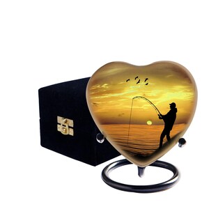 May include: Heart-shaped urn featuring a fishing scene at sunset. The gold-toned urn is mounted on a black metal stand. A black velvet box with a gold clasp is next to the urn. The scene shows a person fishing with birds in the sky.