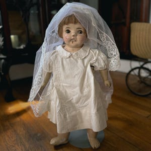 May include: A vintage doll with a painted face, brown hair, and visible wear on the nose and mouth. The doll wears a short-sleeved white dress with a rounded collar and intricate embroidery, paired with a long white lace-trimmed tulle veil.