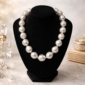 May include: A white pearl necklace displayed on a black jewelry bust. The necklace features large, irregularly shaped pearls strung together. The background includes a perfume bottle, a decorative box, and sparkling accents, suggesting a luxury theme.