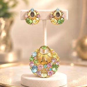 May include: A jewelry set consisting of a pair of gold-tone earrings and a matching circular pendant. Each piece features a central gold-tone metal element surrounded by a cluster of multi-colored faceted crystals in shades of pink, green, and blue.