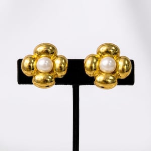 May include: A pair of gold-toned clip-on earrings. Each earring features a four-petal flower design with a faux pearl center. The earrings are displayed on a black velvet surface against a white background.