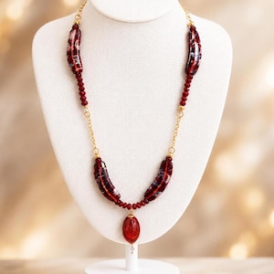 May include: A necklace featuring four curved, dark red glass beads with internal swirling patterns, interspersed with smaller round red beads and gold-toned metal chain links. A single oval red glass pendant hangs from the center of the chain.