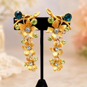 May include: A pair of gold-toned metal drop earrings featuring a cascading floral design. Each earring is adorned with small, light blue and lime green round crystals, with a larger dark teal crystal at the top of the branch-like structure.