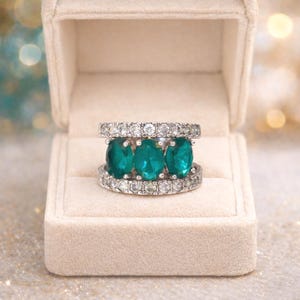 May include: A silver ring with three oval, emerald-green gemstones set between two bands of small, clear stones. The ring is displayed in a beige jewelry box with a soft, velvety interior. The background is blurred with bokeh lights.