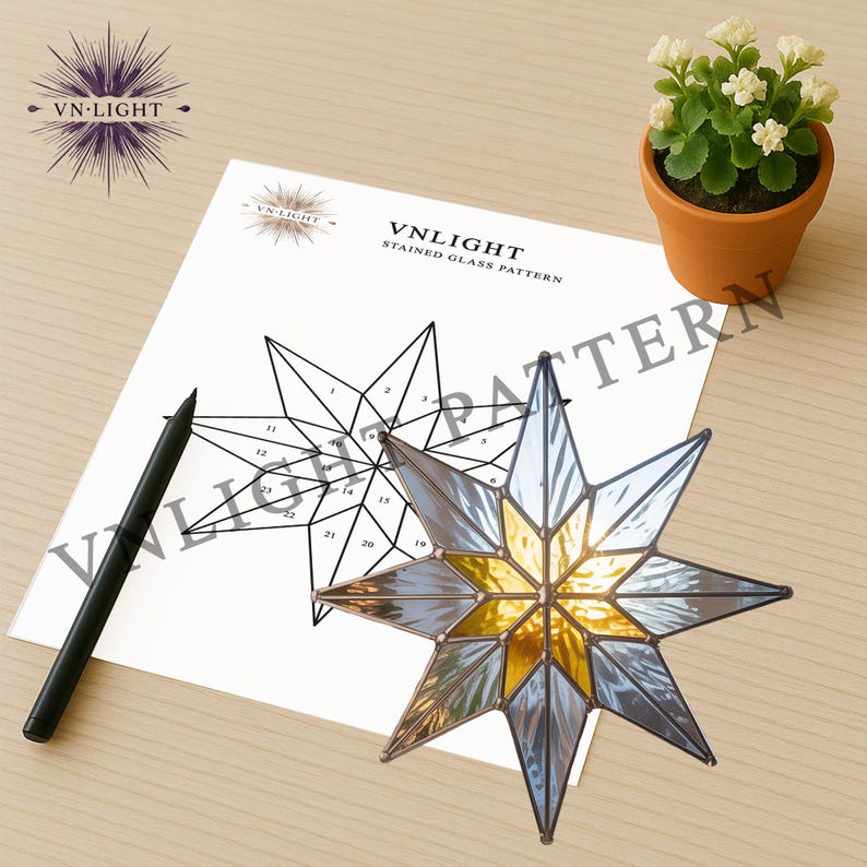 May include: A stained glass star with a gold center and blue-gray points sits next to a pattern diagram. The diagram is labeled "VNLIGHT STAINED GLASS PATTERN". A black pen and a small potted plant are also in the image.