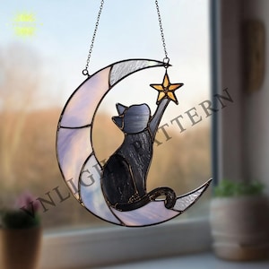 Op de afbeelding: Gebrandschilderde katten silhouet die naar een gouden ster reikt, gezeten op een halve maan. De maan is een mix van wit, grijs en roze glas. De kat is zwart en de ster is goud. Het stuk wordt opgehangen door een ketting.