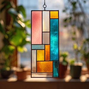 May include: A rectangular stained glass window panel with a black metal frame. The panel features a geometric design with various colored glass pieces, including pink, white, yellow, blue, and orange. The panel is hanging in front of a window.