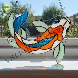 Könnte beinhalten: Bunter Glas-Koi-Fisch-Fensterhänger mit orangefarbenen, blauen, weißen und grünen Segmenten. Der Fisch hängt an einer Kette und wird vor einem Fenster präsentiert. Der Text "VN-LIGHT" ist sichtbar.