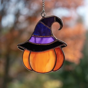 May include: A stained glass Halloween decoration. It features an orange pumpkin with a witch's hat in black, purple, and blue. The pumpkin and hat are outlined in black and suspended by a silver chain. The background is blurred.