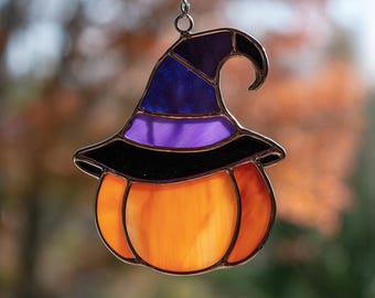 May include: Stained glass Halloween decoration featuring an orange pumpkin with a witch's hat. The hat is black, purple, and blue. The pumpkin and hat are outlined in black and suspended by a silver chain. The background is blurred.