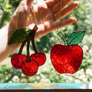 May include: Two stained glass fruit ornaments: a cluster of red cherries and a red apple. Each piece has a gold chain for hanging. The cherries have green leaves, and the apple has a green leaf and stem. The image is set against a blurred background.