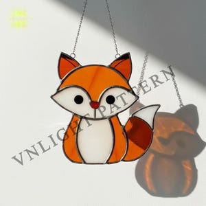 May include: A stained glass fox suncatcher with orange, white, and black details. The fox has a red nose and black eyes, and is hanging from a silver chain. The text "VNIGHT PATTERN" is visible.