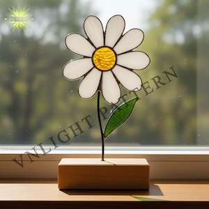 May include: A stained glass daisy with white petals and a yellow center, set on a wooden base. The flower has a green leaf and a black stem. The background is a blurred view of a window.