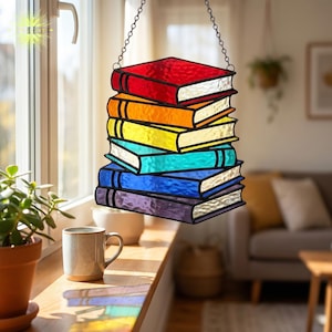 Puede incluir: Adorno de ventana de vidriera con forma de pila de libros. Los libros son de colores del arcoíris: rojo, naranja, amarillo, turquesa, azul y morado. Los libros tienen páginas plateadas y contornos negros.