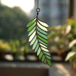 May include: A stained glass leaf suncatcher with a variety of green and white glass pieces. The leaf is outlined in a dark brown metal and is suspended by a thin, dark string. The background is blurred, showing greenery.