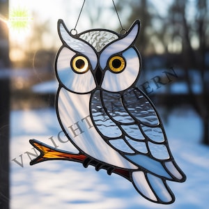 May include: Stained glass owl suncatcher with a white and blue body, perched on a brown branch. The owl has large yellow eyes and is hanging from a black wire. The background is a blurred winter scene.
