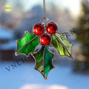 May include: A stained glass holly leaf ornament with three red berries. The ornament is green and red with a gold-coloured metal frame and chain. The text "VN LIGHT" is visible in the upper left corner.