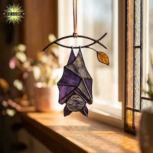 May include: A stained glass bat suncatcher hanging from a metal branch. The bat features purple and grey glass panels with dark metal soldering. A single amber-colored glass leaf is attached to the branch, which is suspended by a thin cord.