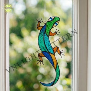 May include: A colourful stained glass lizard suncatcher. The lizard has a green head, turquoise body, and orange and blue details. It hangs in front of a window, with the text "VN LIGHT PATTERN" visible.