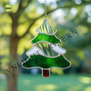 Peut inclure: Ornement de sapin de Noël en vitrail avec une bordure en métal argenté. L'arbre présente des nuances de verre vert et blanc, avec un tronc marron. L'ornement est suspendu devant un arrière-plan vert flou.