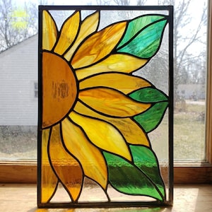 Op de afbeelding: Gebrandschilderd glaspaneel met een zonnebloemontwerp. Het kunstwerk toont een halve zonnebloem met gele en amberkleurige bloemblaadjes, aangevuld met groene bladeren. Het stuk is zwart omlijst en geschikt voor raamdecoratie.